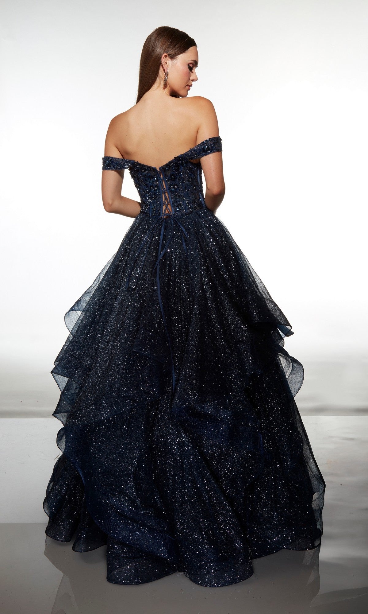 A woman stands facing away in the Alyce 62173 long formal dress, an off-the-shoulder dark blue ball gown with a lace-up back and layered glittery tulle.
